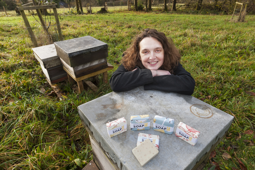 Sarah gates sitting in a field in front of a bee hiveand her amazing soaps on display
